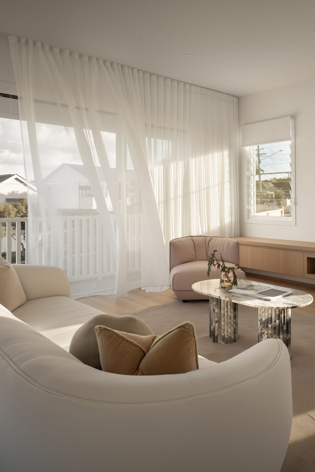 Sunlit living room with floor‑to‑ceiling S‑fold sheer curtains billowing over a sliding door, a curved cream sofa and a marble coffee table, balcony beyond Gold Coast Blinds and Shutters