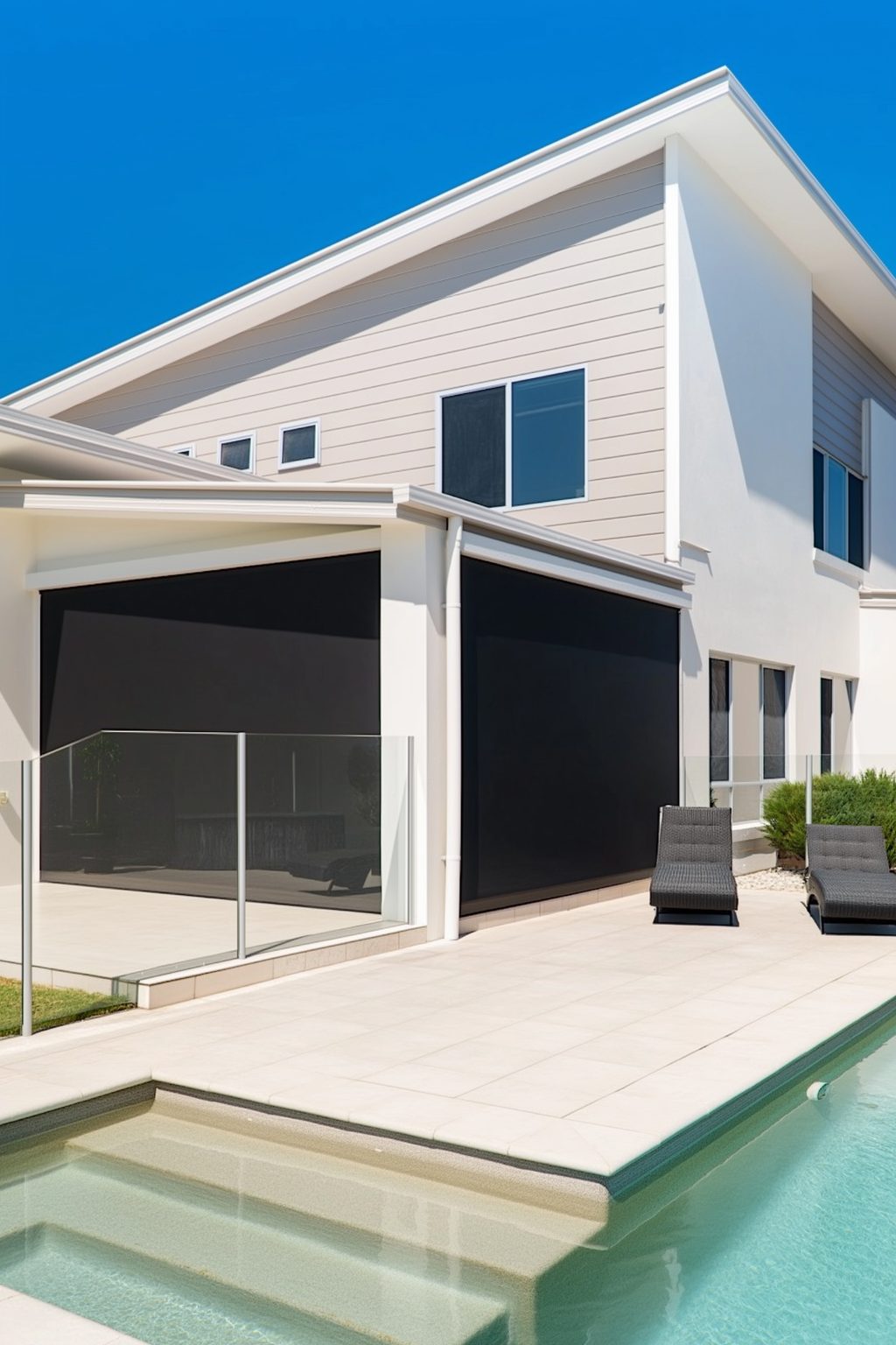Modern two-storey home with black outdoor blinds shading a poolside patio, featuring glass fencing and sun loungers under a clear blue sky.