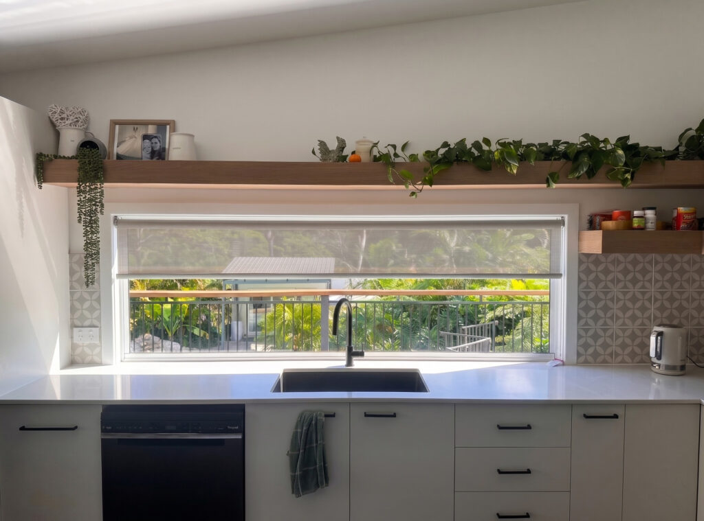Sunlit Gold Coast kitchen with a wide sunscreen roller blind half-lowered over the window above a black sink, the garden visible through the fabric